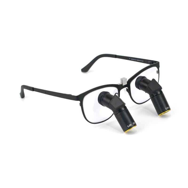 A side-angle product image of Oradenix Ergo TTL Ti Vario dental loupes featuring a sleek black titanium frame. The integrated 5.0X magnification lenses are designed with a downward ergonomic angle to prevent neck strain. The lens barrel clearly shows the "5.0X" marking and the variable focus mechanism, ideal for high-precision endodontic and restorative dental procedures.